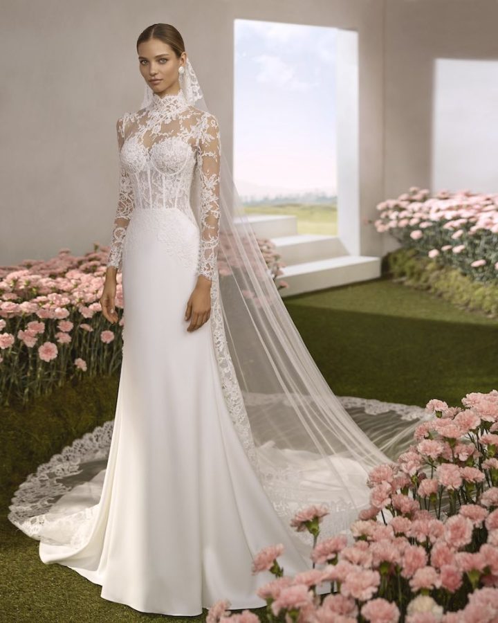 In questa foto, una modella posa indossando un abito della collezione sposa Rosa Clará Couture 2026 con gonna a semi-sirena in georgette, corpetto strutturato con stecche a vista e rivestimento in pizzo con maniche lunghe e collo alto. Indossa anche un velo abbinato