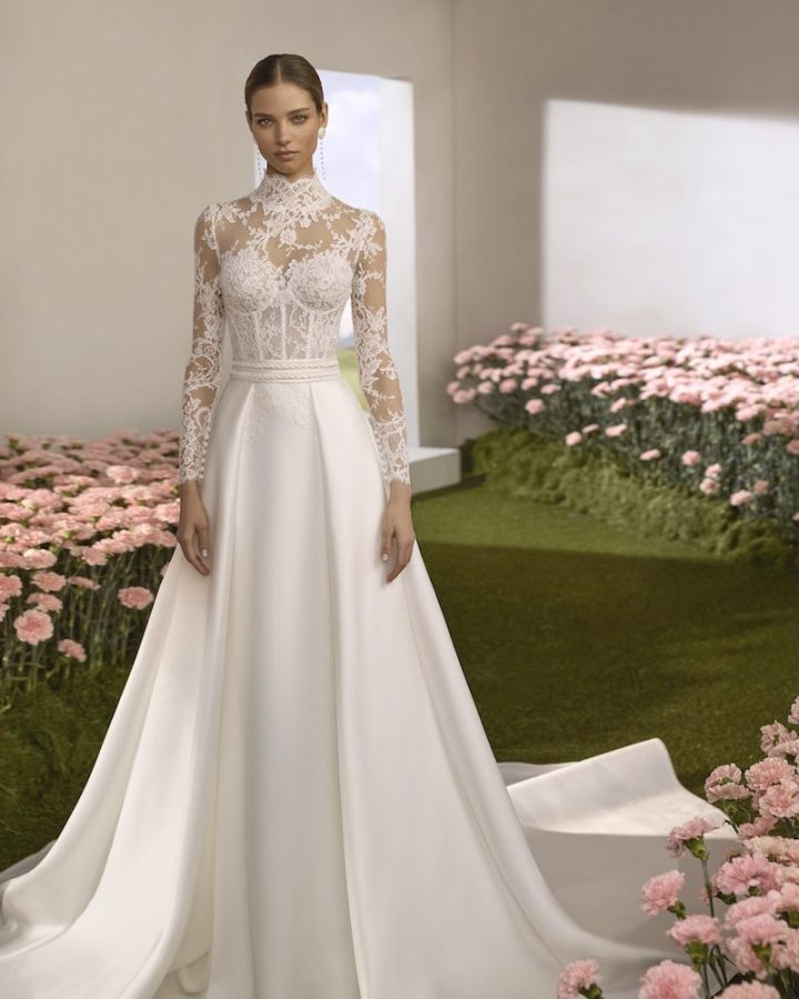In questa foto, una modella posa indossando un abito da sposa Rosa Clará Couture 2026 con corpetto in pizzo con maniche lunghe, corpetto steccato a vista, gonna e sopragonna in mikado