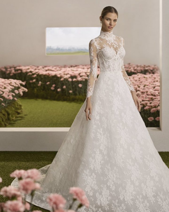 In questa foto, una modella posa indossando un abito da sposa dalla silhouette ad A in pizzo floreale, con corpetto steccato a vista, con maniche lunghe e collo alto