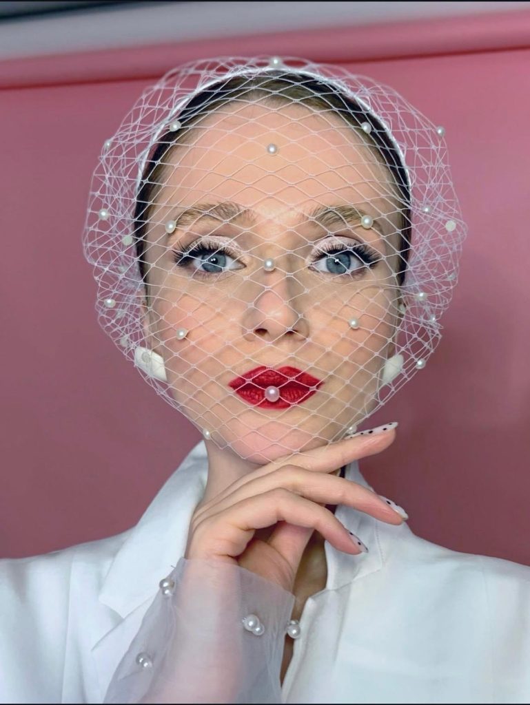 In questa foto, una donna con rossetto rosso acceso posa con un velo moderno decorato da perle, che copre parzialmente il volto. Il make-up è sofisticato, con eyeliner marcato e ciglia in evidenza. La scena suggerisce un approccio glamour e creativo su come scegliere trucco sposa per uno stile contemporaneo.