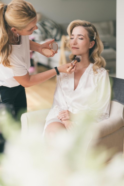 In questa foto, una make-up artist sta applicando il fondotinta con un pennello su una sposa in vestaglia bianca. La sposa guarda dritto davanti a sé, con espressione tranquilla. È un momento di concentrazione e professionalità, ideale per comprendere come scegliere trucco sposa in base al tipo di pelle e al look desiderato.