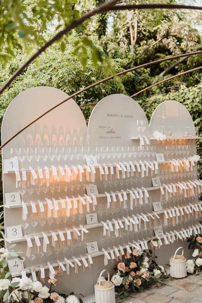 In questa foto, l'allestimento di un tableau de mariage con calici personalizzati con card con i nomi degli ospiti, rose, lanterne e fiori di campo progettato da Giulia Ferro di Maritari
