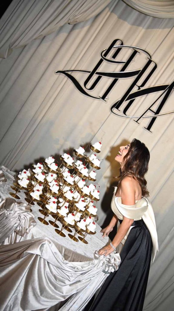 In questa foto, Cira Lombardo davanti ad una torre di coppe di Champagne ripiene di panna e ciliegine davanti ad un drappeggio bianco con su scritto il nome di Cira