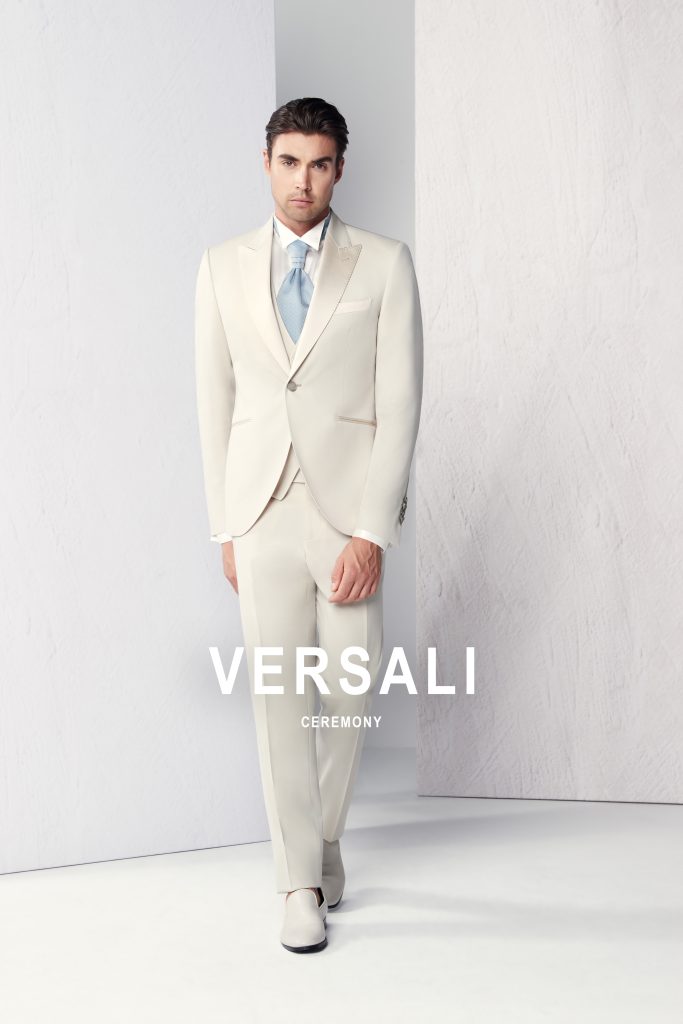 In questa foto, un uomo indossa un elegante completo color avorio con giacca monopetto e cravatta azzurra chiara, posando in piedi davanti a uno sfondo bianco minimale. L'immagine è un esempio perfetto di come scegliere abito da sposo per un matrimonio raffinato e luminoso.