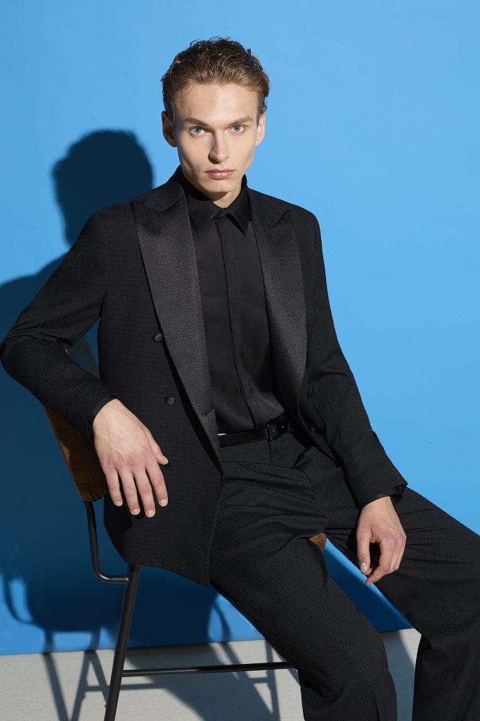 In questa foto, un giovane uomo è seduto su una sedia contro uno sfondo blu acceso, indossa un abito nero con camicia e cravatta ton sur ton. Una proposta moderna e minimalista su come scegliere abito da sposo per chi preferisce il total black.