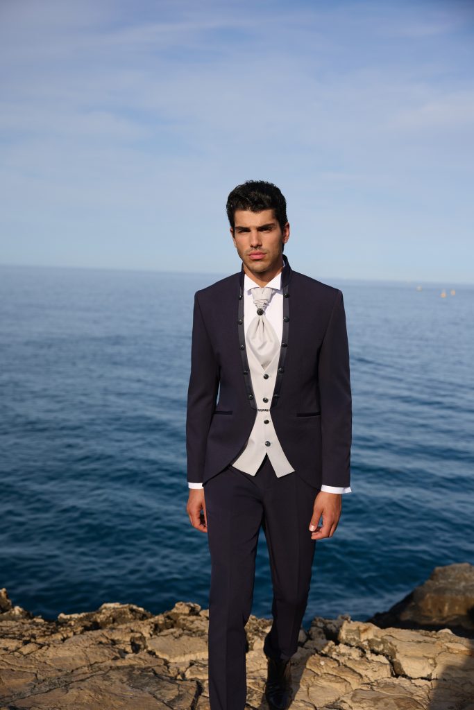 In questa foto lo sposo cammina su una scogliera indossando un abito blu scuro con giacca monopetto, panciotto chiaro a contrasto e cravatta plastron, ideale per comprendere come scegliere abito da sposo per una cerimonia elegante ma luminosa.