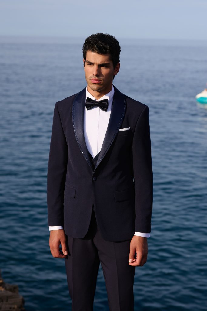 In questa foto un completo blu notte con giacca monopetto, rever satinati e papillon nero è abbinato a camicia bianca e pochette, sullo sfondo del mare: un riferimento classico per come scegliere abito da sposo dallo stile senza tempo.