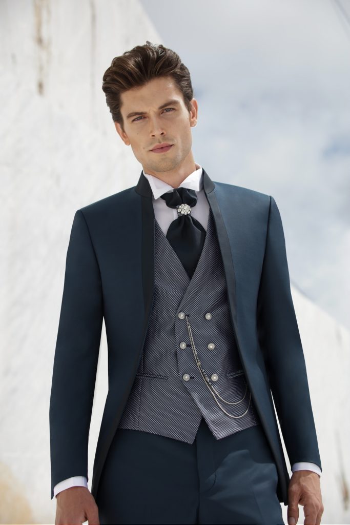 In questa foto uno sposo indossa un abito blu notte con panciotto doppiopetto testurizzato, catena decorativa e cravatta gioiello, un esempio sartoriale per approfondire come scegliere abito da sposo ricercato e scenografico.