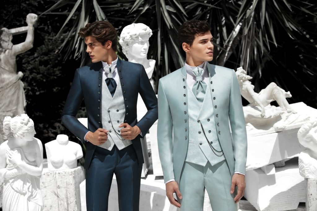 In questa foto due sposi indossano abiti coordinati nei toni del blu e dell’azzurro polvere, con giacche monopetto e panciotti decorati, mostrando varianti cromatiche utili per capire come scegliere abito da sposo in base allo stile personale.