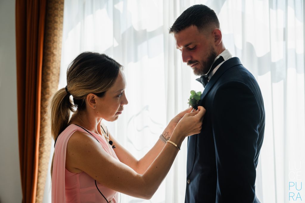 In questa foto una donna sistema il fiore all’occhiello sulla giacca blu scuro dello sposo, che indossa camicia bianca e papillon, in una stanza illuminata dalla luce naturale: un momento preparatorio che racconta come scegliere abito da sposo anche attraverso gli accessori.