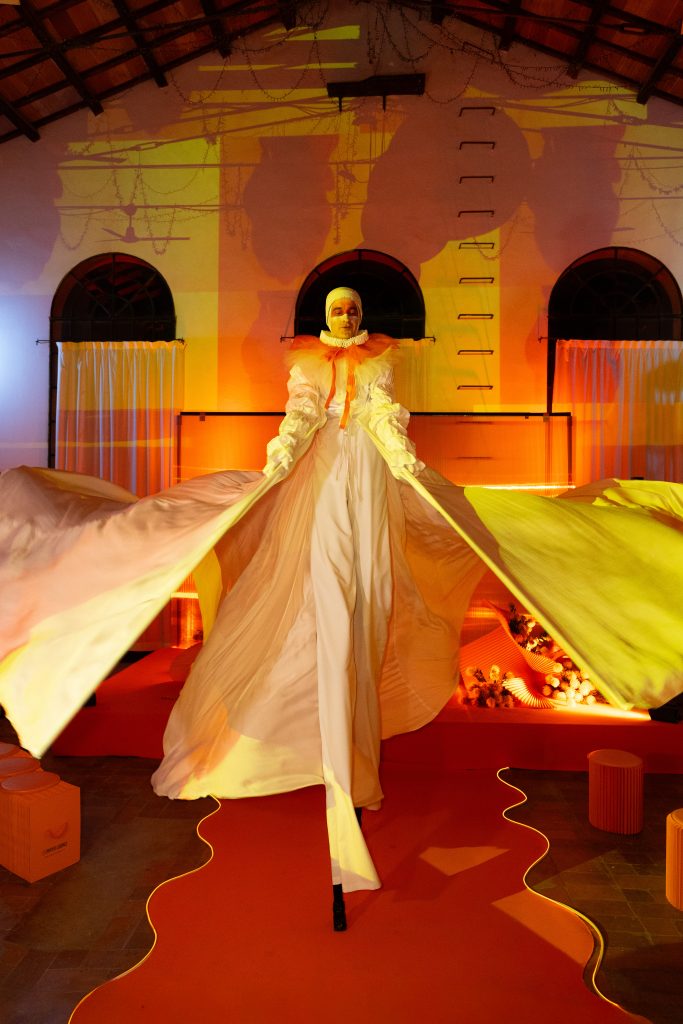 In questa foto, un performer su trampoli domina la scena centrale dell’Unframed Networking Cocktail Experience. Indossa un abito bianco con maniche larghe e dettagli arancioni, in posa teatrale su un tappeto arancio sagomato che accentua la scenografia suggestiva.