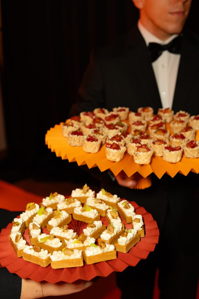 In questa foto, due camerieri in smoking nero servono finger food raffinato su vassoi colorati e geometrici. I vassoi, a tema con l’Unframed Networking Cocktail Experience, ospitano mini tartellette creando una mise en place originale e di design.