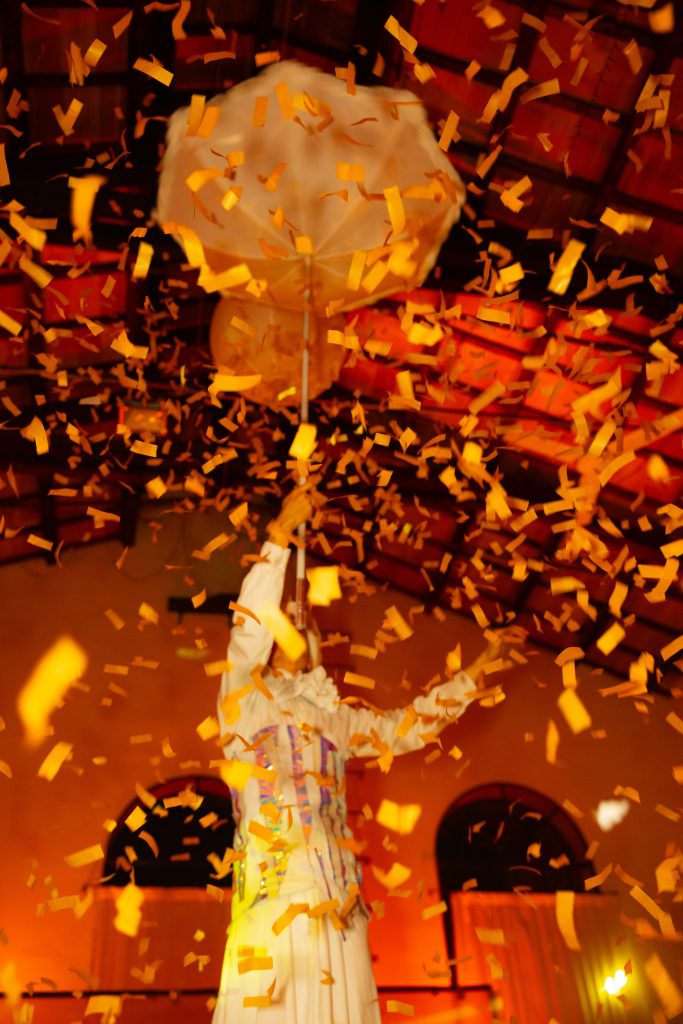 In questa foto, coriandoli arancioni riempiono l’aria mentre una figura artistica solleva un grande ombrello bianco. La scena, parte dell’Unframed Networking Cocktail Experience, è vibrante e teatrale, con un gioco di luci che enfatizza la gioia del momento.