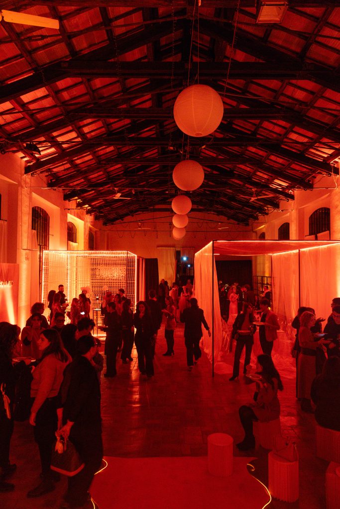 In questa foto, l’ambiente industriale dell’Unframed Networking Cocktail Experience è pieno di persone in movimento. Il soffitto alto in legno e le installazioni luminose rosse creano un’atmosfera avvolgente. A sinistra, si intravedono spazi espositivi e aree di networking.