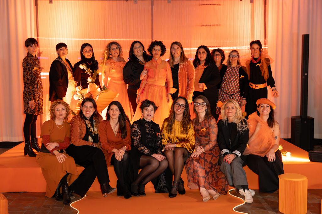 In questa foto, un gruppo di donne sorridenti posa sul palco dell’Unframed Networking Cocktail Experience, vestite in tonalità calde come arancione, ruggine e nero. L’atmosfera è conviviale, professionale e celebrativa, in un contesto allestito con luci calde e scenografie floreali.