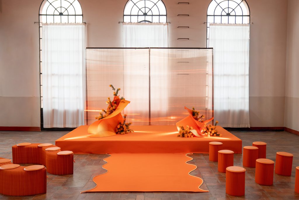 In questa foto, lo spazio dell’Unframed Networking Cocktail Experience è mostrato nella sua versione diurna, con colori arancio brillante. Le strutture sinuose e i fiori gialli creano un ambiente elegante e moderno, con sedute tonde arancioni e un tappeto centrale sagomato che guida verso il palco.