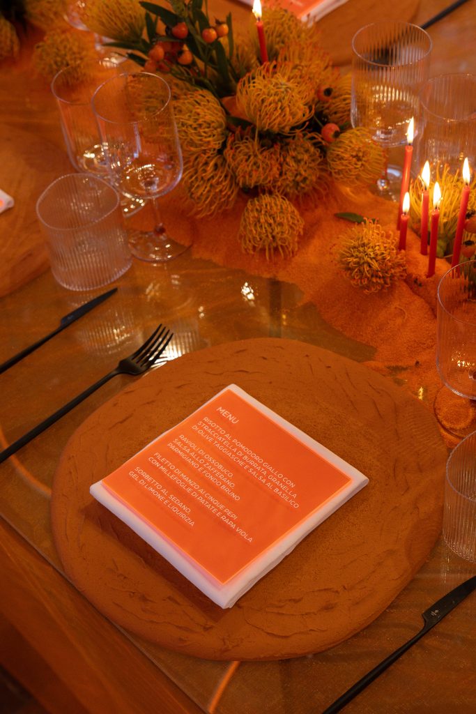 In questa foto, una mise en place elegante dell’Unframed Networking Cocktail Experience mostra un tavolo decorato con fiori giallo-arancio e candele accese. Al centro, un menu arancione con scritte bianche elenca piatti ricercati, circondato da calici trasparenti e posate nere.