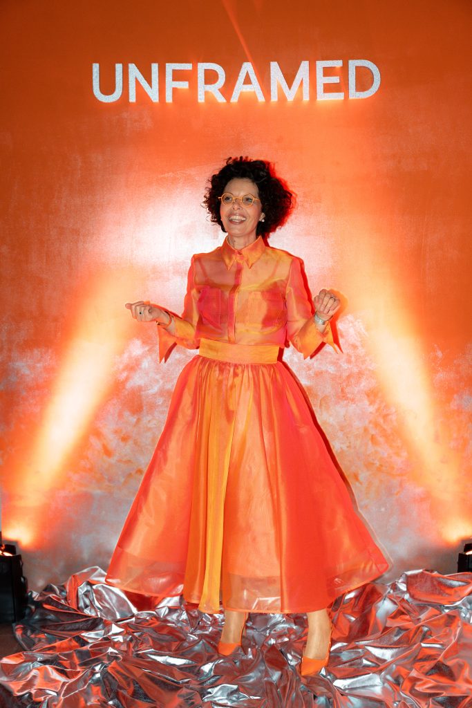 In questa foto Maria Mayer, una donna con capelli ricci, indossa un completo arancione e sta in piedi davanti a un muro illuminato con la scritta “UNFRAMED”. L’immagine cattura lo spirito potente dell’Unframed Networking Cocktail Experience, tra colore, design e celebrazione.