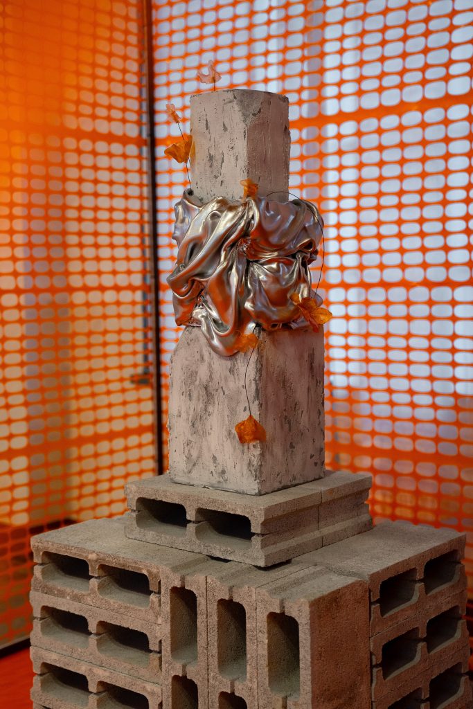 In questa foto, una torta si trasforma in installazione artistica dell’Unframed Networking Cocktail Experience. La torta è costituita da un blocco che ricrea il cemento decorato con drappeggi argento e piccoli elementi arancioni. La base è fatta di mattoni forati, e lo sfondo è una rete da cantiere arancio acceso.