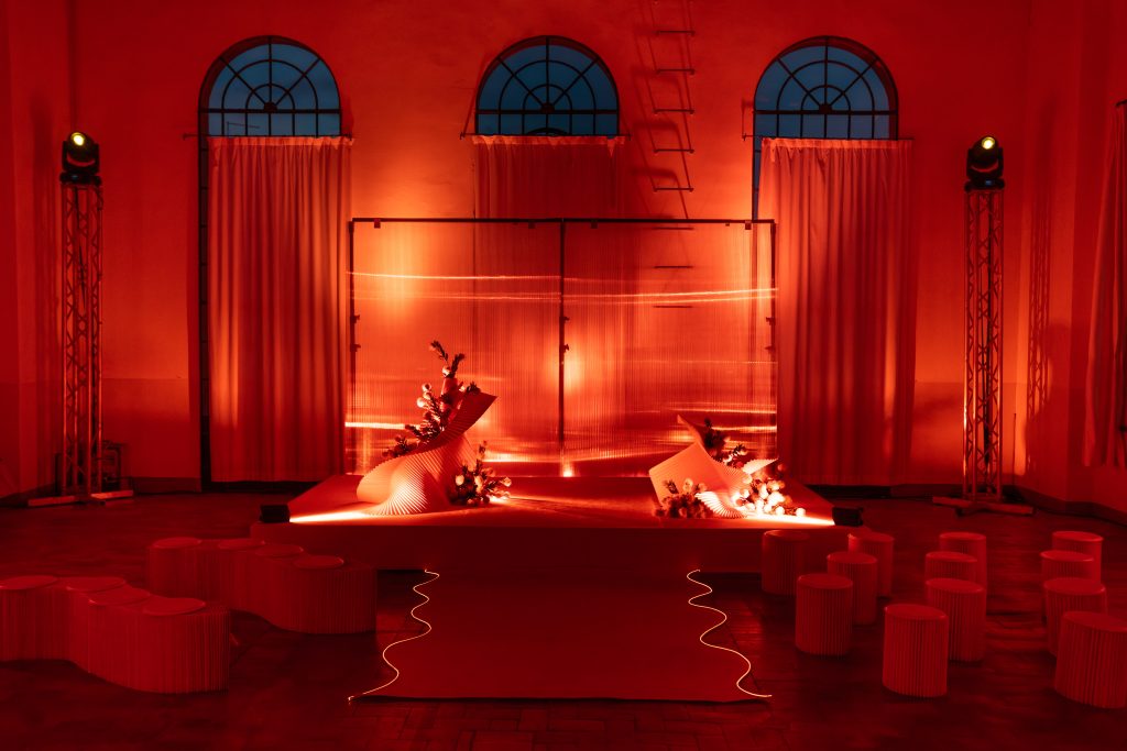 In questa foto, lo spazio di dell’Unframed Networking Cocktail Experience è immerso in una luce rossa intensa che avvolge l’intero allestimento scenografico Al centro c’è una pedana illuminata, decorata con elementi floreali e strutture curve in materiale rigido. L’atmosfera è teatrale e avvolgente.