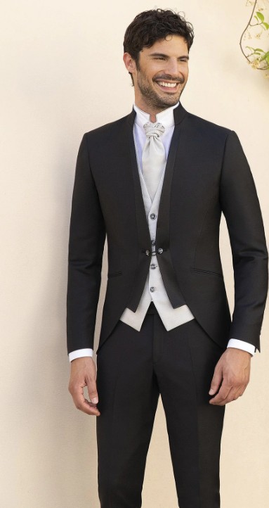 In questa foto, un uomo sorridente indossa un abito da cerimonia nero con gilet grigio chiaro e cravattone, ideale per un look classico e sofisticato. Un ottimo esempio di come scegliere abito da sposo se si ama l’eleganza tradizionale con un tocco di originalità.