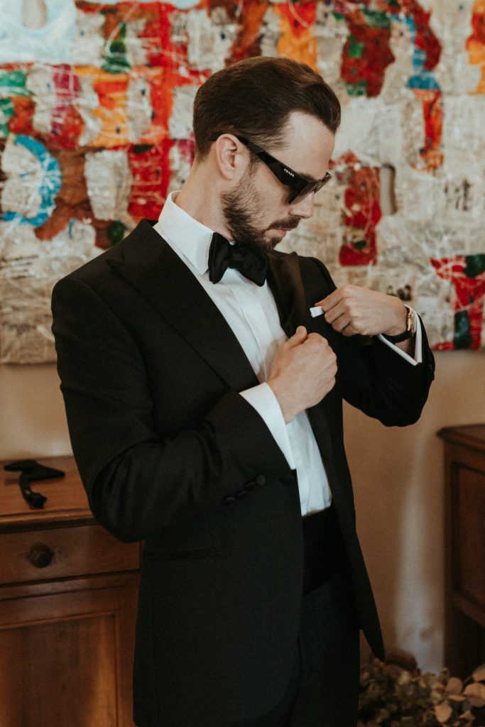 In questa foto uno sposo indossa uno smoking nero classico con camicia bianca e papillon nero, mentre sistema il taschino della giacca in un ambiente interno dal fondo artistico colorato, un esempio elegante per capire come scegliere abito da sposo formale e senza tempo.