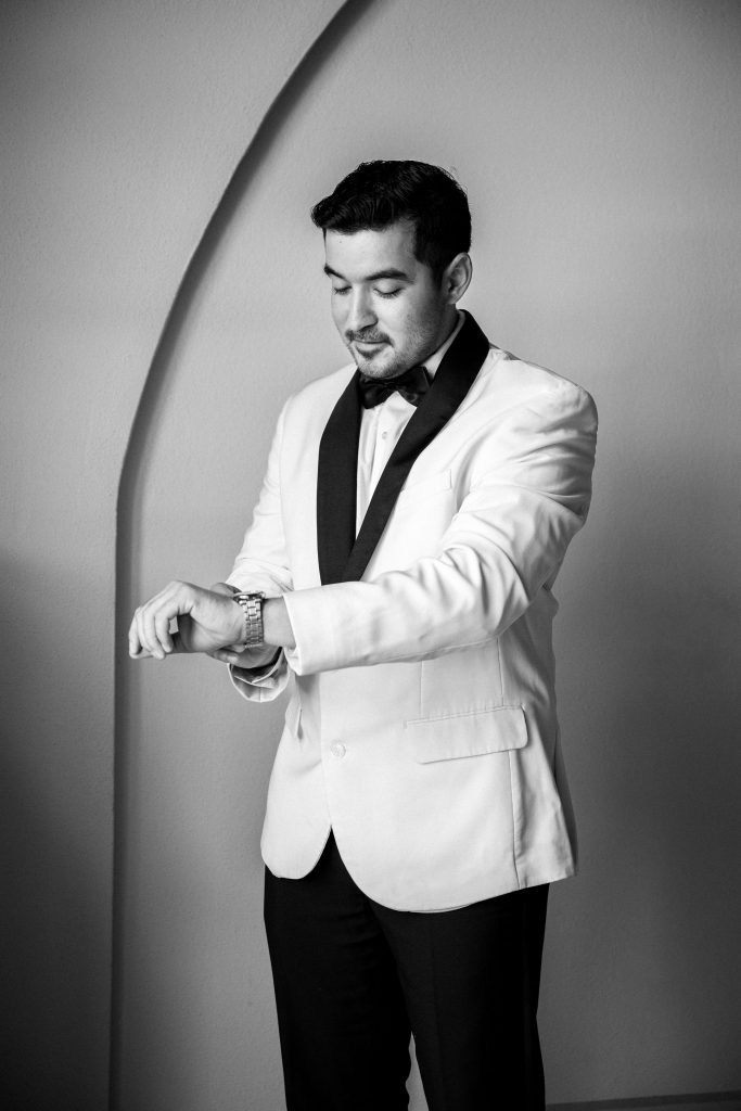 In questa foto uno sposo in bianco e nero indossa uno smoking bianco con rever scuri a contrasto, camicia bianca e papillon nero, mentre controlla l’orologio al polso: un’immagine raffinata per orientarsi su come scegliere abito da sposo per un look contemporaneo e ricercato.