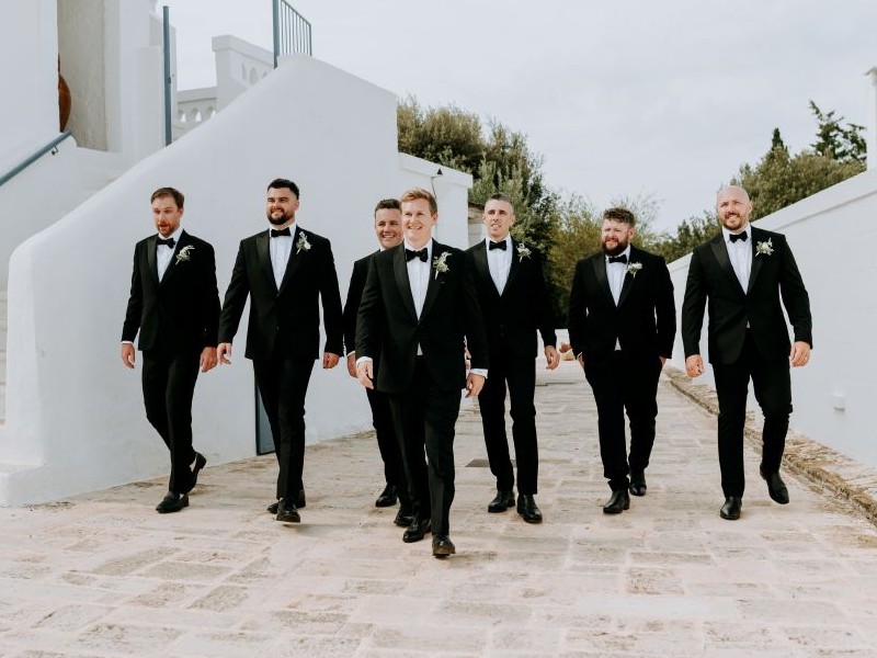 In questa foto, un gruppo di uomini in smoking nero con papillon cammina allโaperto in un contesto mediterraneo. La scena mostra diverse opzioni coordinate su come scegliere abito da sposo per il protagonista e i suoi testimoni.