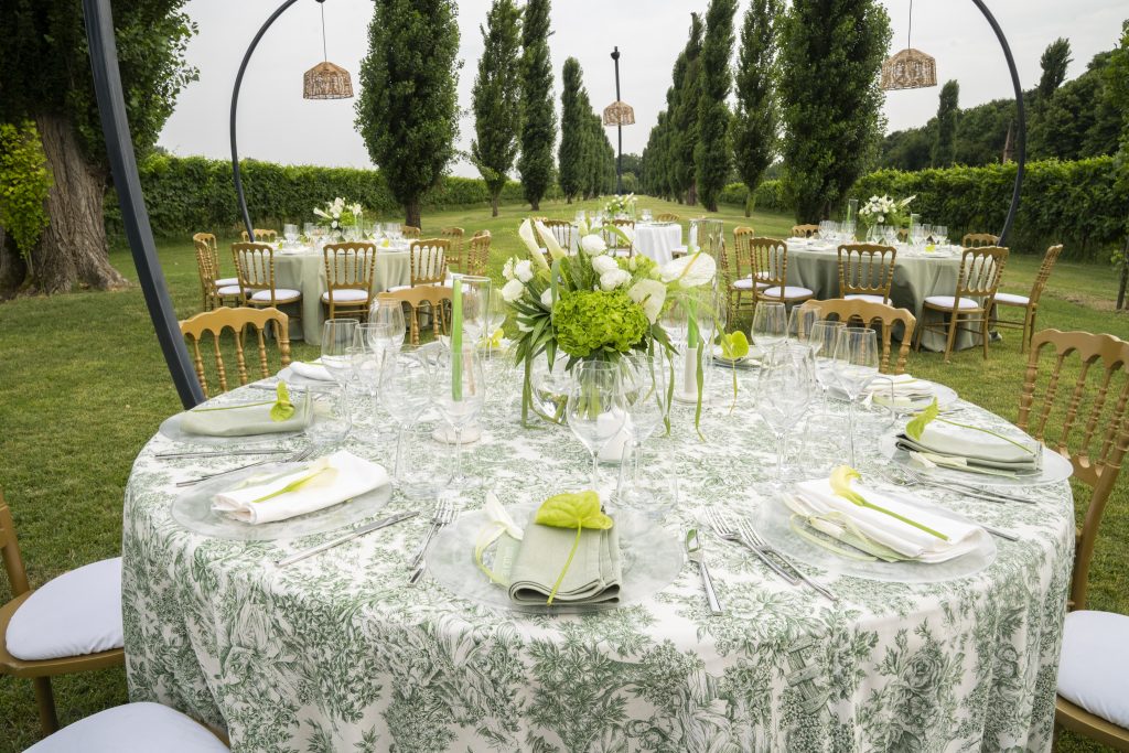 In questa foto, un’elegante tavola rotonda è apparecchiata all’aperto in un giardino verdeggiante, con una mise en place 2026 raffinata e armoniosa. La tovaglia decorata con motivi botanici in verde salvia fa da sfondo a piatti in vetro trasparente, tovaglioli verdi e bianchi con fiocchi in raso e piccoli dettagli floreali. Al centro, un bouquet di ortensie verdi e calle bianche aggiunge freschezza alla composizione, tra calici cristallini e posate in acciaio lucido. Sullo sfondo, altre tavole coordinate completano l’atmosfera naturale e sofisticata.