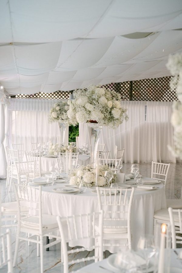 In questa foto, la sala è decorata interamente in bianco, con tavoli rotondi, sedie in stile chiavarina e fiori bianchi in composizioni voluminose su alzate trasparenti. Le tovaglie e i piatti sono candidi, per un effetto etereo e pulito. L’immagine rappresenta una delle tendenze mise en place 2026 più amate per i matrimoni eleganti e senza tempo: l’all white minimalista.