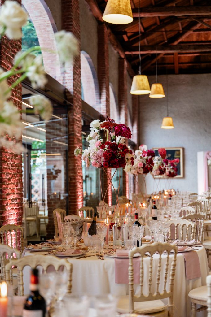 In questa foto, una lunga sala con pareti in mattoni e soffitto in legno ospita tavoli tondi decorati con composizioni floreali verticali in tonalità rosso e rosa. I tovaglioli rosa chiaro contrastano con il bianco delle tovaglie. Le tendenze mise en place 2026 uniscono romanticismo e verticalità per donare movimento e volume allo spazio.