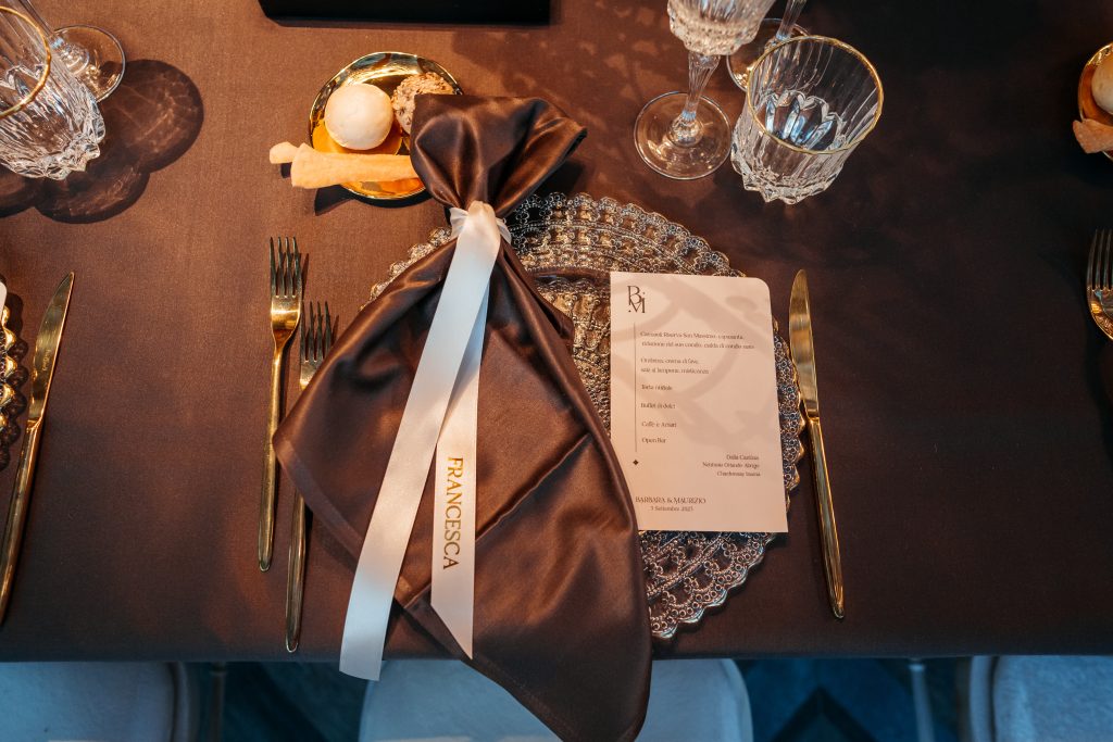 In questa foto, un’elegante mise en place su tovaglia scura presenta un piatto in vetro decorato, posate dorate, calici lavorati e un tovagliolo in raso marrone chiuso da un nastro bianco con nome stampato. Le tendenze mise en place 2026 esplorano il lusso personalizzato con palette intense e texture ricercate.