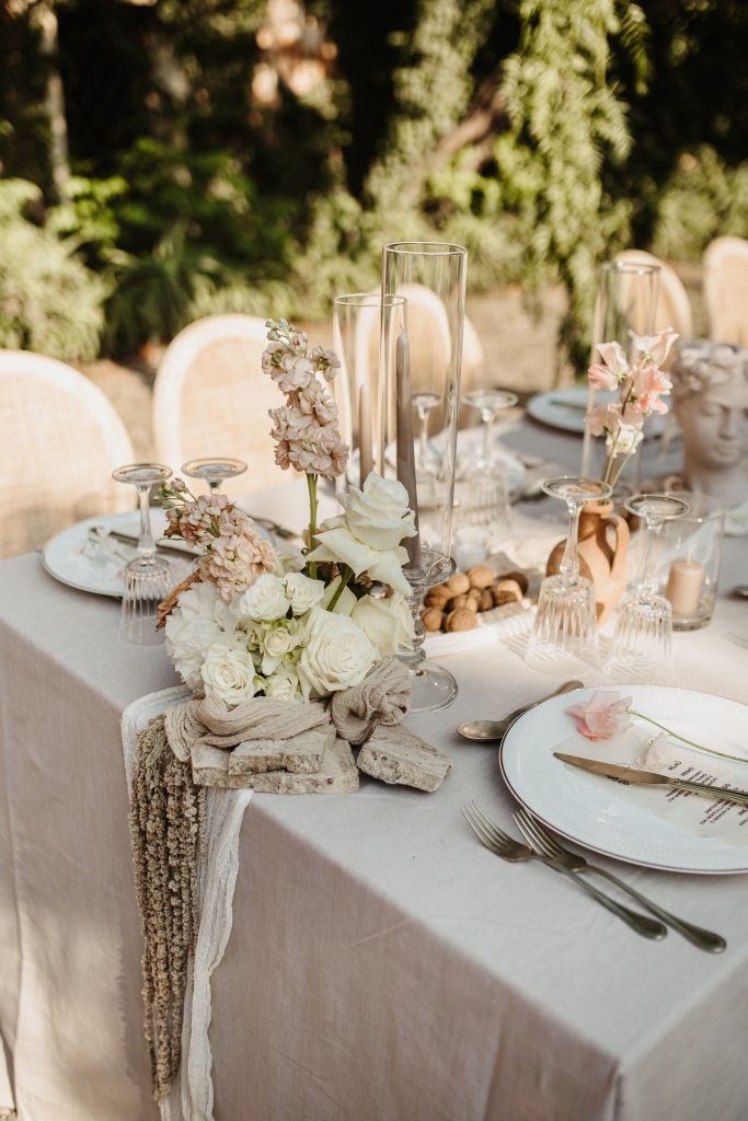 In questa foto, un tavolo rettangolare all’aperto è apparecchiato con tovaglia chiara, piatti bianchi e fiori freschi nei toni crema e cipria. Elementi naturali come noci e vasi in pietra completano la scena. Le tendenze mise en place 2026 celebrano l’autenticità con materiali grezzi e palette neutre.