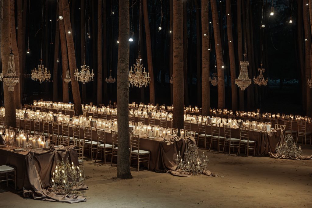 In questa foto suggestiva, i tavoli sono immersi in un bosco illuminato da candele e lampadari in stile chandelier appesi tra gli alberi. Le tovaglie in tessuto scuro creano un contrasto raffinato con la luce calda delle candele. La scena è intima e teatrale, rappresentando una delle tendenze mise en place 2026 più scenografiche, perfetta per chi desidera un matrimonio nel cuore della natura, ma con un tocco di glamour.
