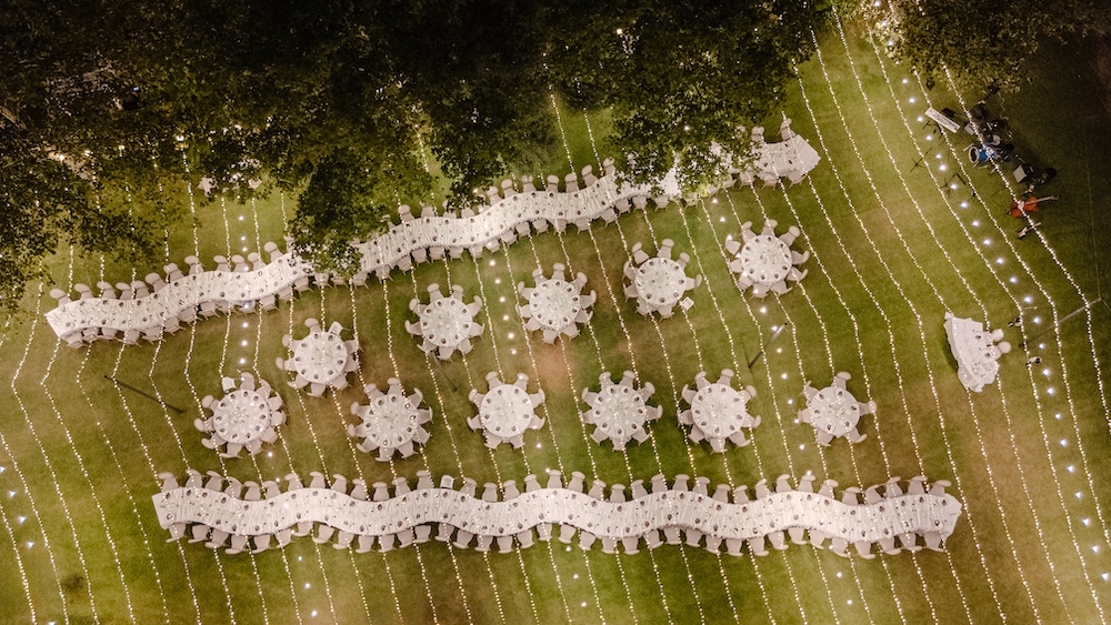 In questa foto, scattata dall’alto, si vedono tavoli lunghi e rotondi disposti in modo armonioso su un prato, circondati da fili di luci che creano un effetto magico. L’intero spazio è avvolto da un’atmosfera fiabesca, perfetta per una cena all’aperto sotto le stelle. Questo stile celebra le tendenze mise en place 2026 che prediligono la scenografia immersiva e l’uso creativo della luce.