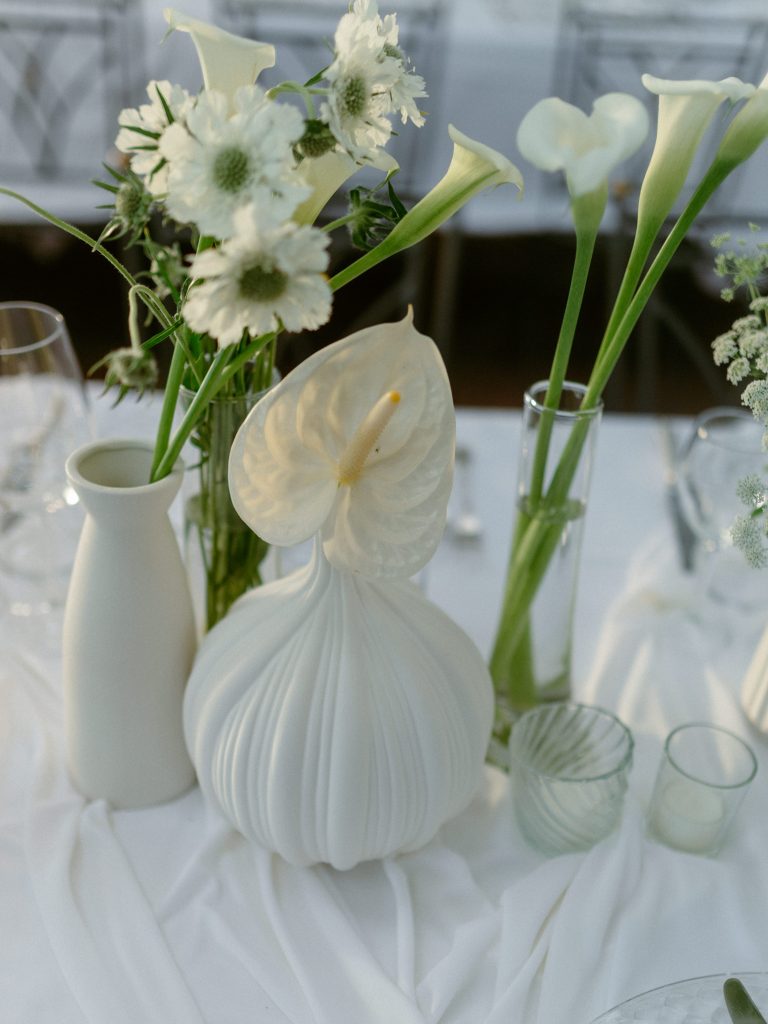 In questa foto, un centrotavola minimalista celebra l’eleganza monocromatica: vasi in ceramica bianca opaca e vetro trasparente accolgono fiori bianchi come calle, scabiose e anturium. La composizione rispecchia le tendenze allestimenti matrimonio 2026 ispirate alla purezza, alla leggerezza e a un’estetica senza tempo.