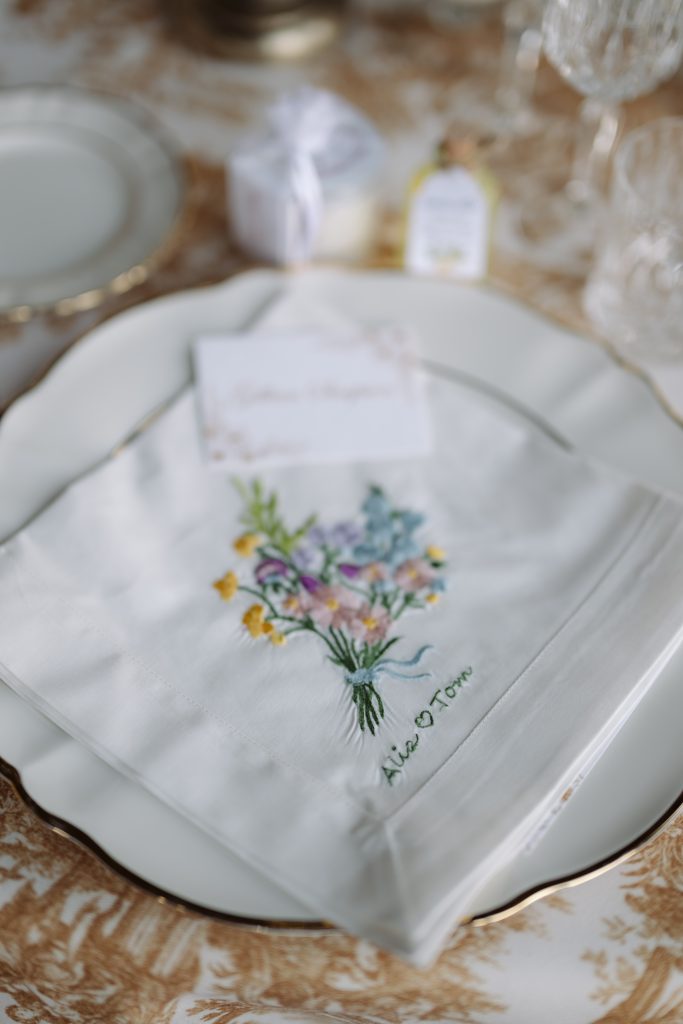 In questa foto, un tovagliolo bianco personalizzato con un ricamo floreale color pastello e i nomi degli sposi, posato su un piatto in porcellana bordato oro. Questo dettaglio su misura riflette una delle tendenze allestimenti matrimonio 2026 più raffinate: il ritorno al fatto a mano e ai dettagli artigianali personalizzati.