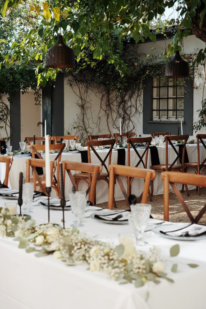 In questa foto, un allestimento matrimonio in stile rustic chic mostra lunghe tavolate all’aperto, con sedie in legno naturale, tovaglie bianche e candelabri neri minimalisti. L’atmosfera è intima e immersa nel verde, con lampade in rattan che pendono tra i rami. Una scelta perfetta per chi cerca ispirazione tra le tendenze allestimenti matrimonio 2026 più essenziali e naturali.