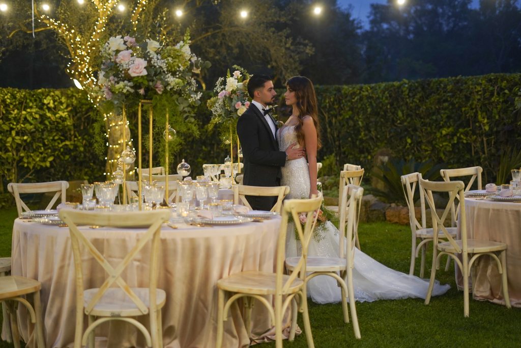 In questa foto, gli sposi si tengono per mano in un giardino incantato, tra tavoli rotondi vestiti di rosa cipria, centrotavola con fiori pastello e luci sospese. Il mood è romantico e senza tempo, perfetto per chi cerca un’eleganza raffinata. Un'ispirazione delicata che spicca tra le tendenze allestimenti matrimonio 2026 dal gusto classico ma luminoso.