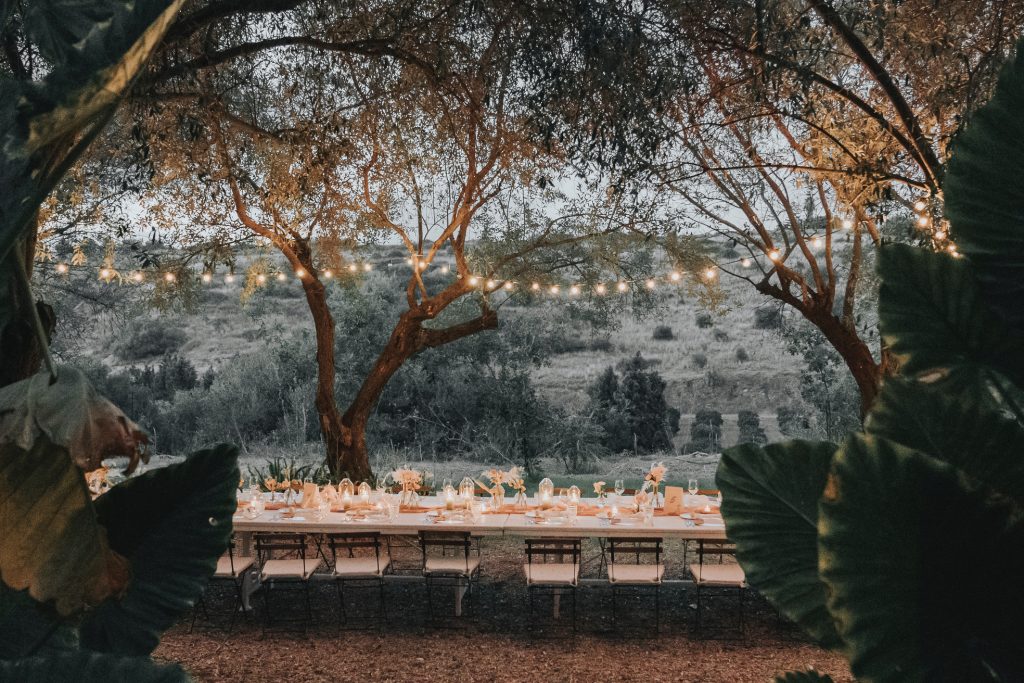 In questa foto, una lunga tavolata è allestita tra gli alberi con un gioco di luci calde sospese tra i rami. I dettagli rustici si fondono con il romanticismo di fiori chiari e candele. Un’ambientazione intima e boho, ideale per chi cerca tra le tendenze allestimenti matrimonio 2026 soluzioni suggestive in mezzo alla natura.