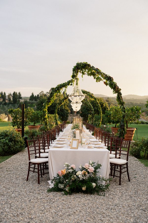 In questa foto, uno scenografico tavolo imperiale è allestito su un viale di ghiaia circondato dal verde, con una maestosa struttura ad archi ricoperta di rampicanti e grandi lampadari di cristallo sospesi al centro. Il design sofisticato e naturalistico riflette perfettamente le Tendenze allestimenti matrimonio 2026, che valorizzano la connessione con la natura, l’eleganza senza tempo e l’armonia tra elementi botanici e dettagli luxury.