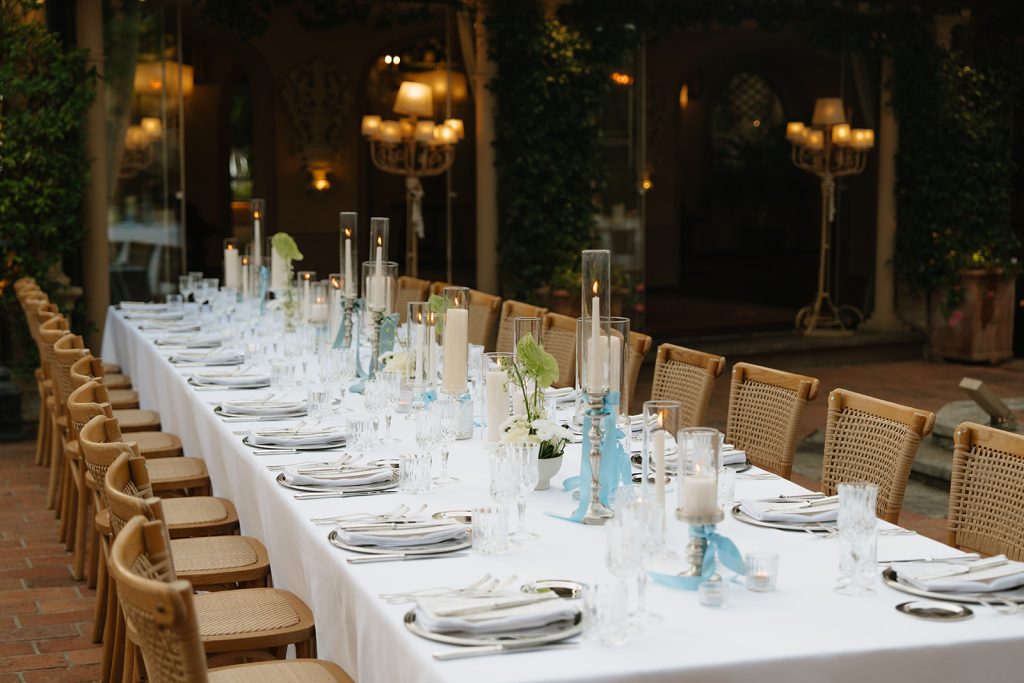 In questa foto, un tavolo allestito in una corte storica unisce candele alte, portacandele azzurri e cristalleria raffinata su una tovaglia bianca, circondato da sedute in rattan. L'equilibrio tra dettagli vintage e palette soft è emblematico delle tendenze allestimenti matrimonio 2026 in stile italian chic.