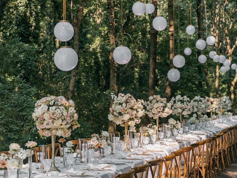 In questa foto, un allestimento matrimonio in stile rustic chic mostra lunghe tavolate all’aperto, con sedie in legno naturale, tovaglie bianche e candelabri neri minimalisti. L’atmosfera è intima e immersa nel verde, con lampade in rattan che pendono tra i rami. Una scelta perfetta per chi cerca ispirazione tra le tendenze allestimenti matrimonio 2026 più essenziali e naturali.