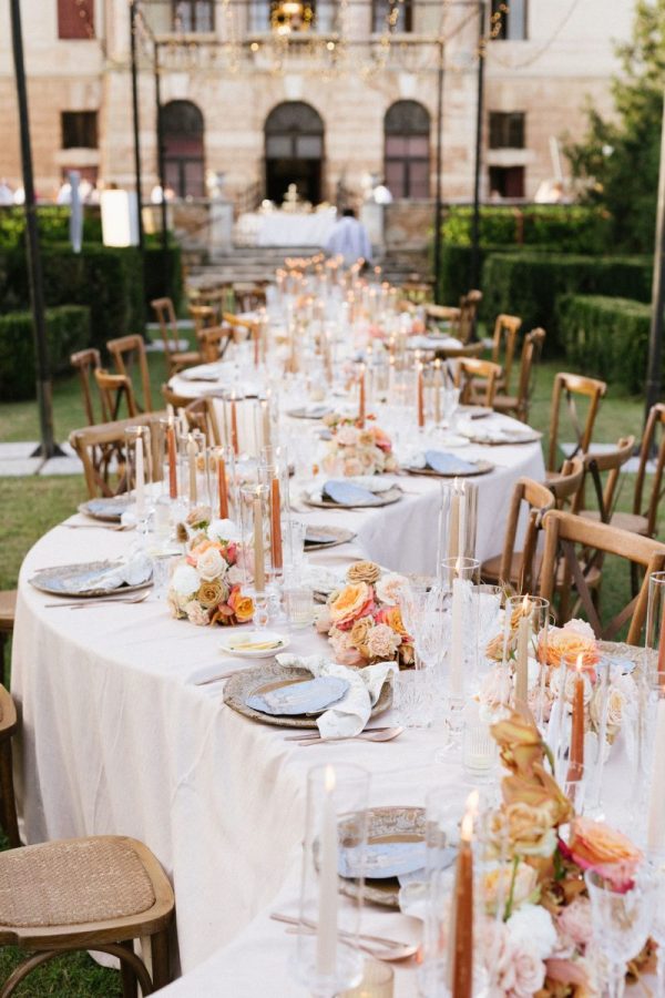 In questa foto, un lungo tavolo ovale apparecchiato all’aperto presenta piatti decorati, candele sottili color bronzo e composizioni di rose pesca e avorio. Il contesto è un’elegante villa d’epoca, con un’atmosfera romantica e sofisticata. Rappresenta le tendenze allestimenti matrimonio 2026 che fondono heritage, raffinatezza e colore.