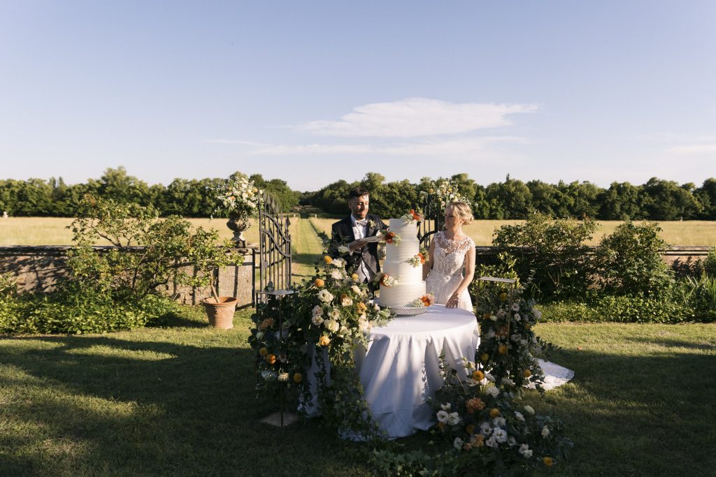 In questa foto, una coppia di sposi taglia una torta nuziale altissima decorata con fiori freschi, disposta su un tavolo circolare immerso in un giardino. L’allestimento bucolico e curato esprime pienamente le tendenze allestimenti matrimonio 2026 che esaltano la natura e i suoi colori autentici.
