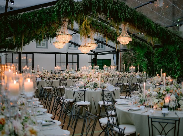 In questa foto, tavoli rotondi sono disposti sotto una grande struttura trasparente con lampadari e decorazioni verdi sospese. I centrotavola floreali sono ricchi ma equilibrati, con candele lunghe che aggiungono calore alla scena. Questo allestimento interpreta alla perfezione le tendenze mise en place 2026, che sposano il verde botanico con l’eleganza delle serre in vetro.