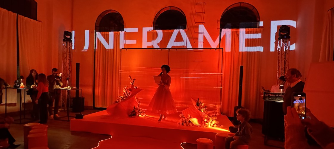 In questa foto, lo spazio è immerso in una luce rossa intensa che avvolge l’intero allestimento scenografico dell’Unframed Networking Cocktail Experience. Al centro c’è una pedana illuminata, decorata con elementi floreali e strutture curve in materiale rigido, sullo sfondo tende bianche e archi a vetro. L’atmosfera è teatrale e avvolgente. Marya Mayer è in piedi sul palco mentre parla