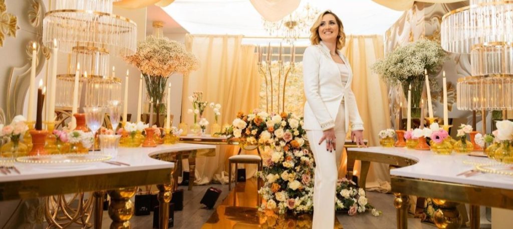 In questa foto si vede uno degli allestimenti de Le Farfalle in Fiore – Angela Crupi La wedding designer posa sorridente tra i tavoli elegantemente decorati per un ricevimento. Indossa un completo bianco e si trova al centro di una sala sontuosa, avvolta da tendaggi crema e bouquet di fiori freschi in tonalità pesca, crema e rosa cipria. I tavoli sono impreziositi da candelabri in vetro, alzate dorate e mise en place raffinate. L’allestimento trasmette un senso di armonia, classe e cura per ogni dettaglio.