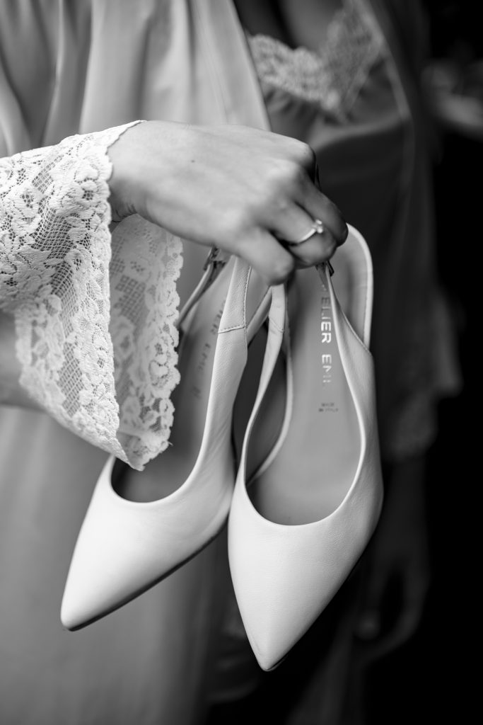 In questa foto si vede una proposta di scarpe da sposa 2026, un paio di décolleté bianche a punta affusolata, tenute in mano dalla sposa, con linee essenziali e finitura opaca che richiamano un’eleganza minimal.