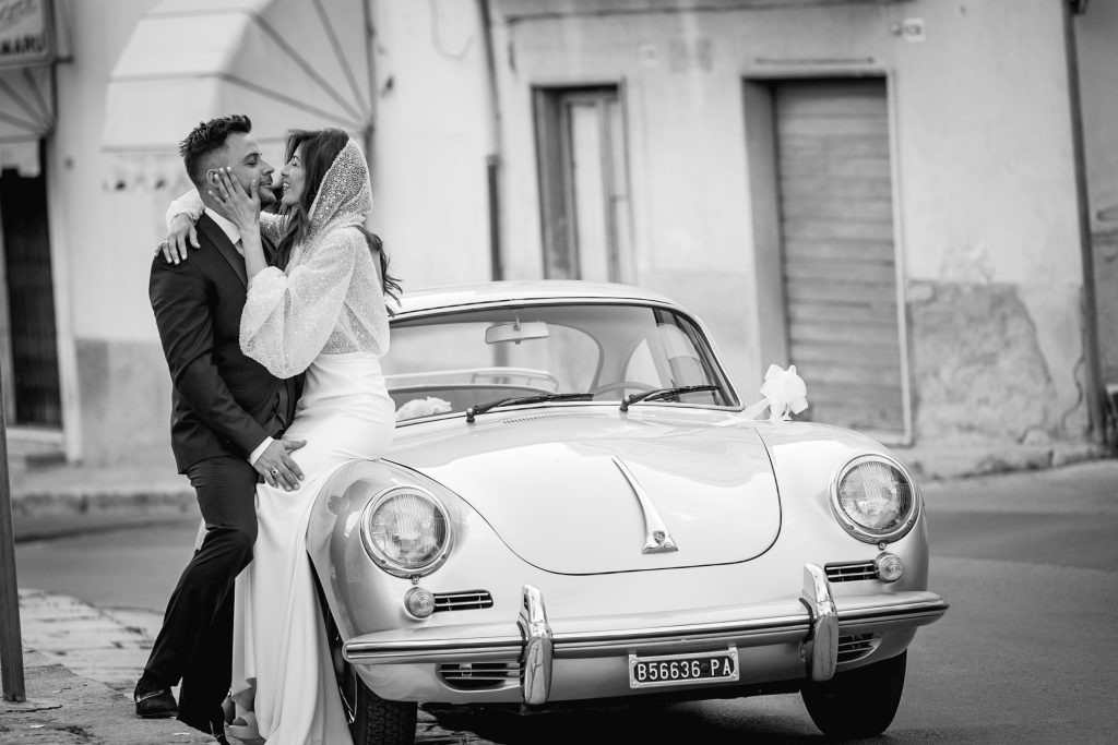 In questa foto si vede una delle fotografie di matrimonio di Davide Meli, una coppia di sposi in bianco e nero appoggiata a un’auto d’epoca, con la sposa seduta sul cofano e lo sposo in piedi accanto a lei.